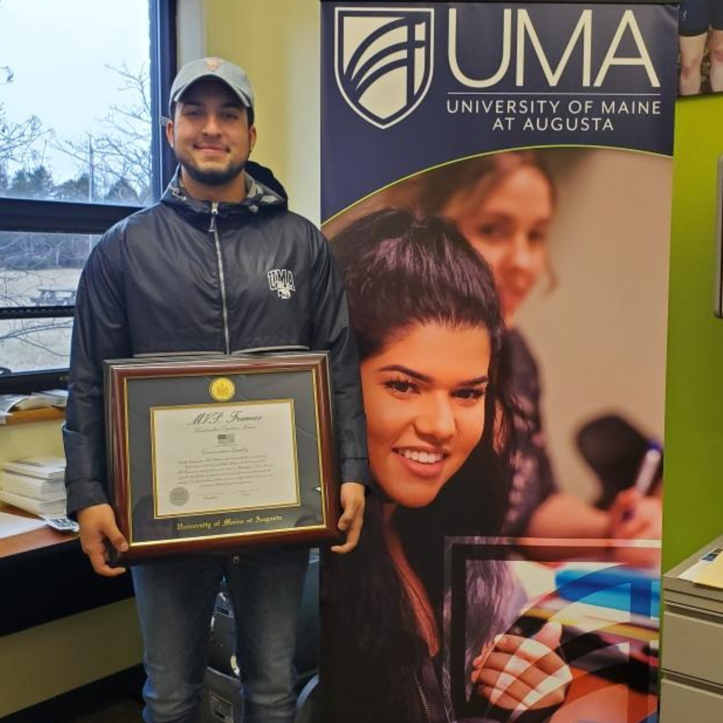 Noah Thompson holding a degree diploma standing next to a UMA sign 