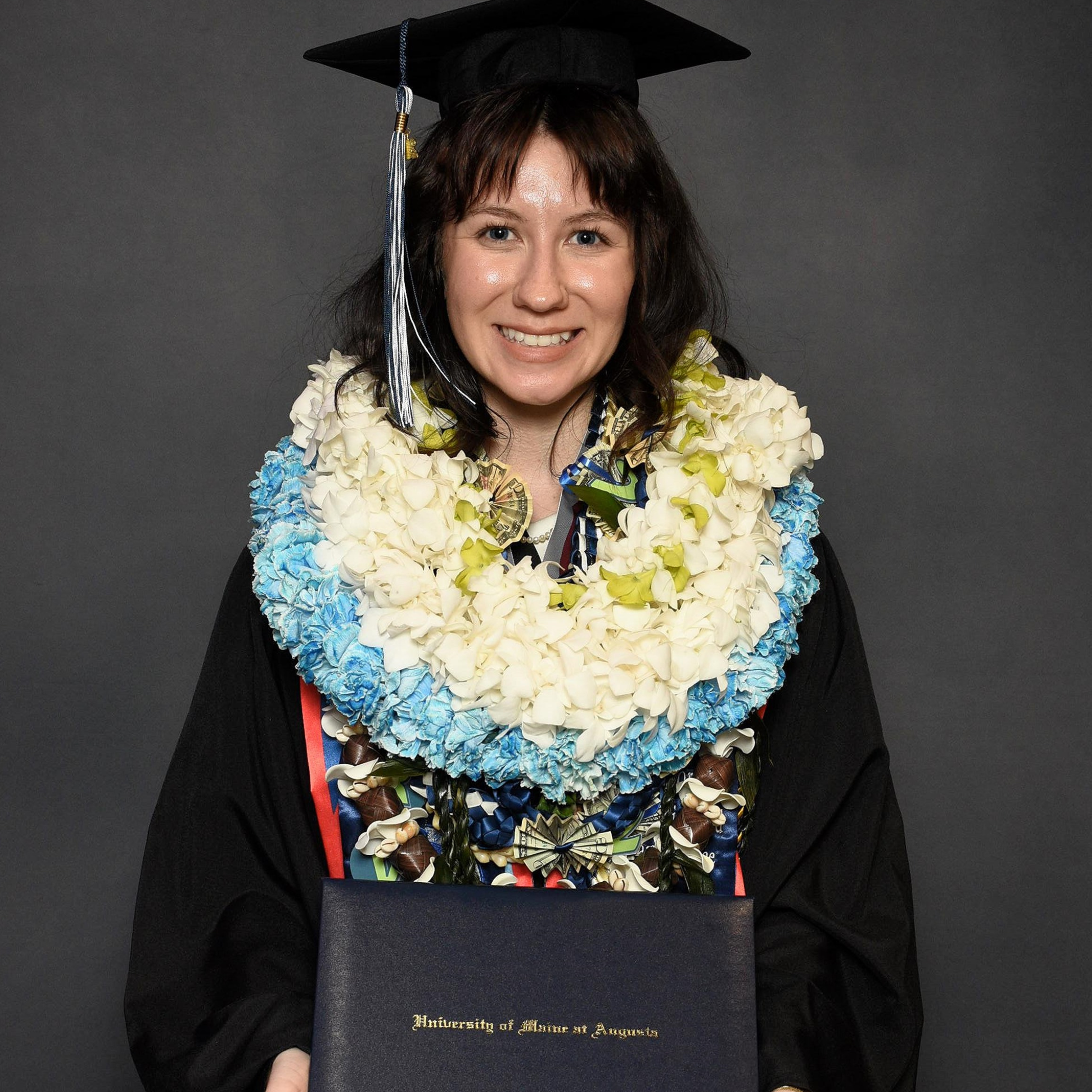 Garvey, wearing a graduation cap and gown, smiles while holding a University of Maine at Augusta diploma cover. She is adorned with several large floral leis in white, blue, and green, along with colorful cords and stoles against a dark gray backdrop.