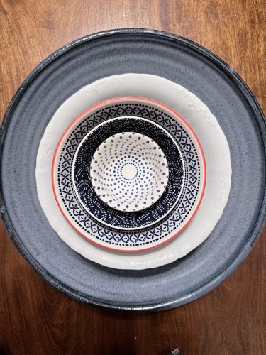 A stack of four decorative plates and bowls of varying sizes and patterns, arranged on a wooden surface. The largest plate is a matte blue, followed by a white plate with a textured edge. The next plate is white with a coral rim and a detailed navy blue geometric pattern. The innermost item is a small bowl with a white interior covered in a swirling pattern of blue dots.