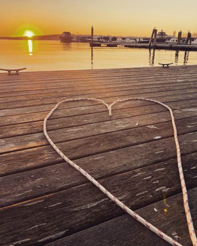 A white rope forms a heart shape on a weathered wooden pier at sunset, with a calm harbor and boats in the background.