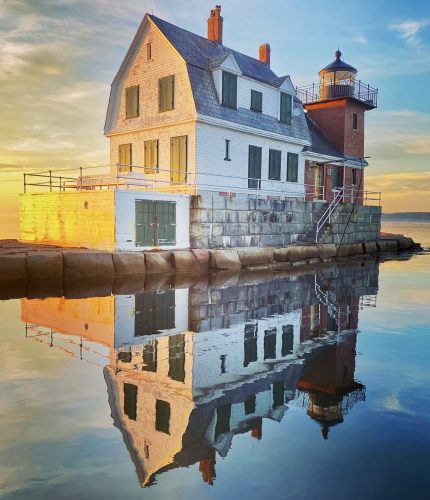 A white lighthouse keeper's house with green shutters stands on a stone foundation beside a red brick tower with a lantern room, reflected in calm water under a golden and blue sky at sunrise.