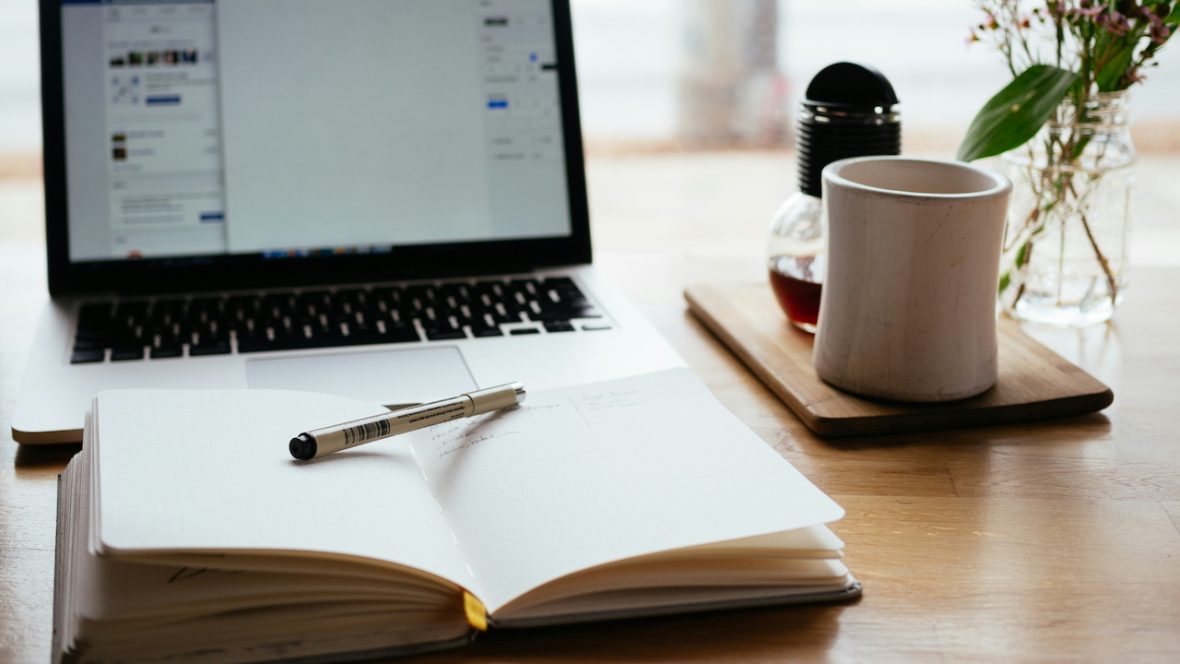 a notebook, laptop, pen, and mug sitting on a desk