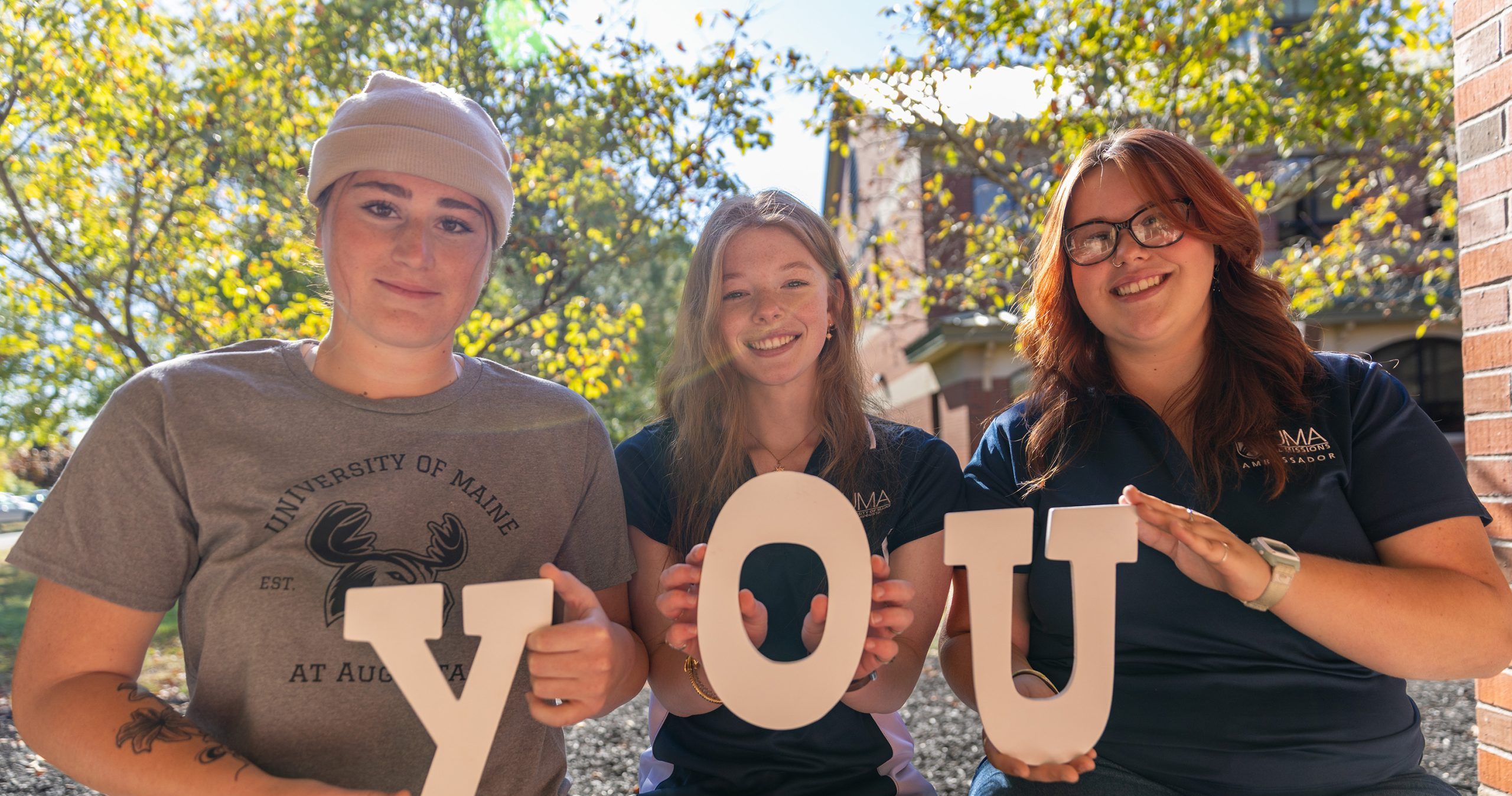 Three UMA students sit outside on a sunny day, smiling and holding large white letters that spell “YOU.” The student on the left wears a gray University of Maine at Augusta T-shirt and a beige beanie, while the two on the right wear navy UMA polo shirts. Trees and a brick building are visible in the background.