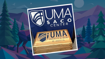 A wooden time capsule box engraved with the University of Maine at Augusta (UMA) logo and name sits in front of a blue banner that reads “UMA Saco Center.”