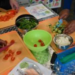 Three people chopping fresh vegetables—including cherry tomatoes, spinach, cucumbers, and onions—on orange cutting boards during a cooking activity. Metal bowls and utensils are arranged on a silver textured table surface.