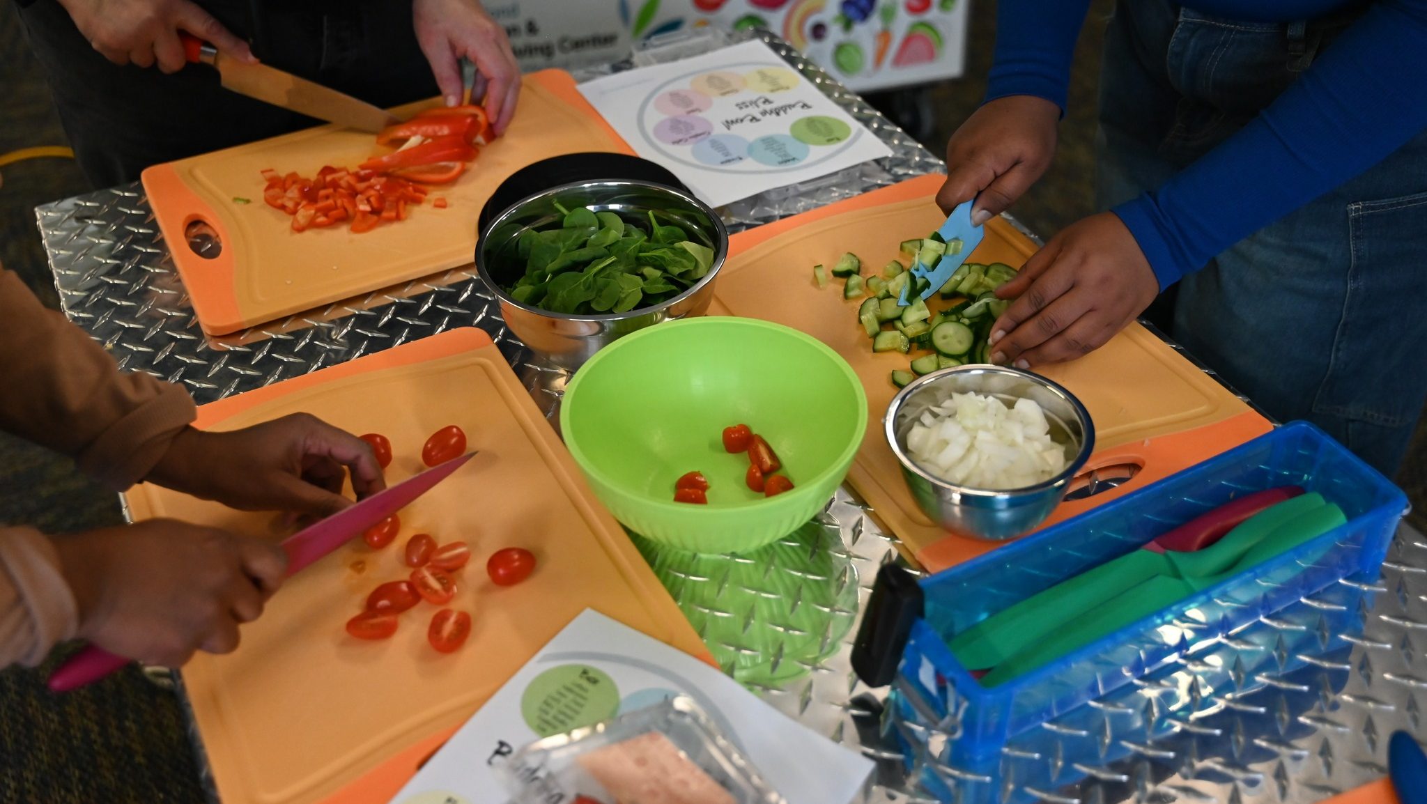 Three people chopping fresh vegetables—including cherry tomatoes, spinach, cucumbers, and onions—on orange cutting boards during a cooking activity. Metal bowls and utensils are arranged on a silver textured table surface.