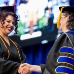 A graduate in cap and gown smiles while shaking hands with a faculty member on stage during UMA's 2024 commencement ceremony.
