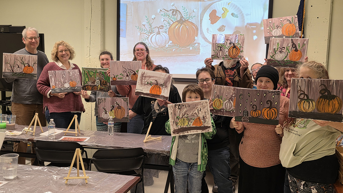 UMA Rumford Paint Night participants proudly display their finished pumpkin paintings in front of the classroom projector screen. The group smiles while holding up colorful fall-themed artwork.