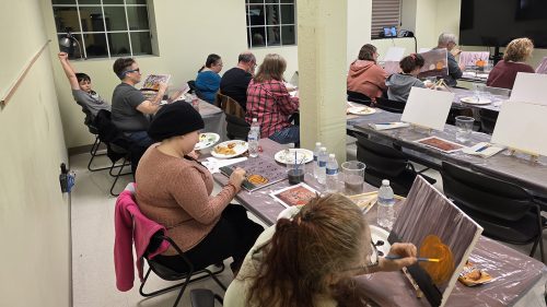 Participants at UMA Rumford’s Paint Night sit at long tables covered with painting supplies and snacks, working on canvas paintings featuring pumpkins. 