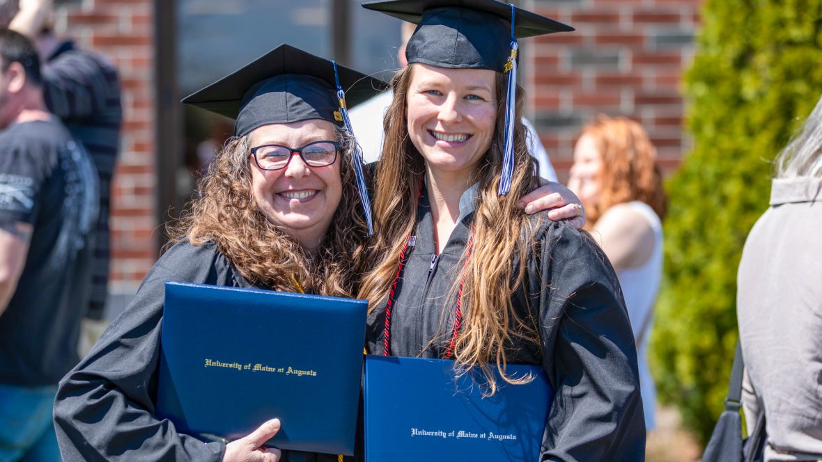 Two University of Maine at Augusta graduates in caps and gowns smile and pose together outdoors, each holding their diploma covers after commencement.