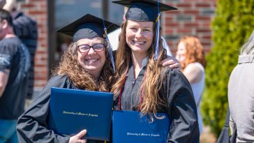 Two University of Maine at Augusta graduates in caps and gowns smile and pose together outdoors, each holding their diploma covers after commencement.