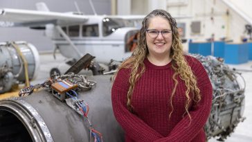 Maria Wilcox, manager of UMA's aviation maintenance technician school, with long, blonde, curly hair and glasses, smiles while standing in front of an airplane engine inside of an airplane hangar.