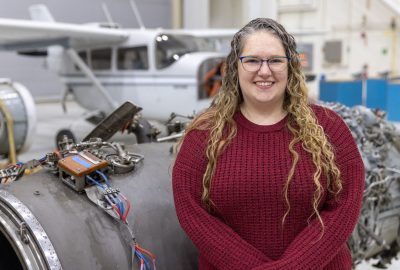 Maria Wilcox, manager of UMA's aviation maintenance technician school, with long, blonde, curly hair and glasses, smiles while standing in front of an airplane engine inside of an airplane hangar.