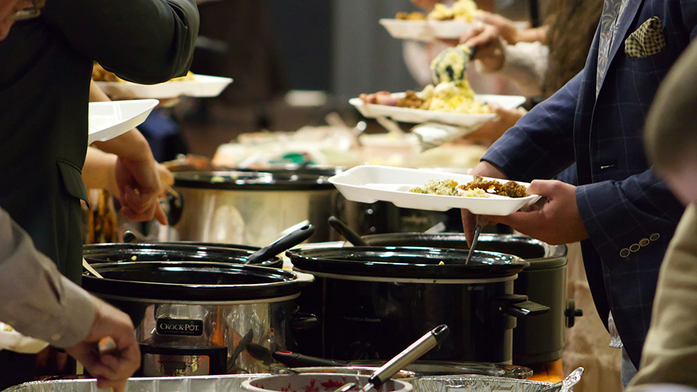 A close-up of slow cookers on a long table with people serving themselves food.