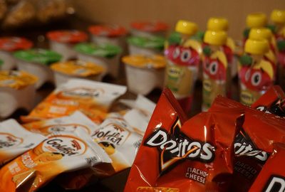Close-up of assorted snack items on a table, including individual bags of Doritos and Goldfish crackers, fruit pouches with yellow caps, and small yogurt cups in assorted flavors.
