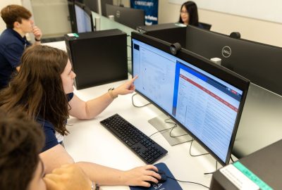 Students seated at desktop computers in a computer lab review cybersecurity data on large monitors during a collaborative class session.