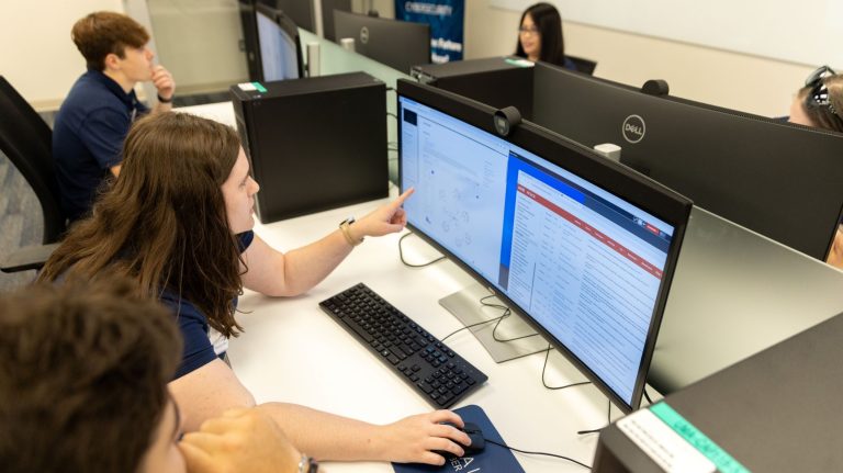 Students seated at desktop computers in a computer lab review cybersecurity data on large monitors during a collaborative class session.