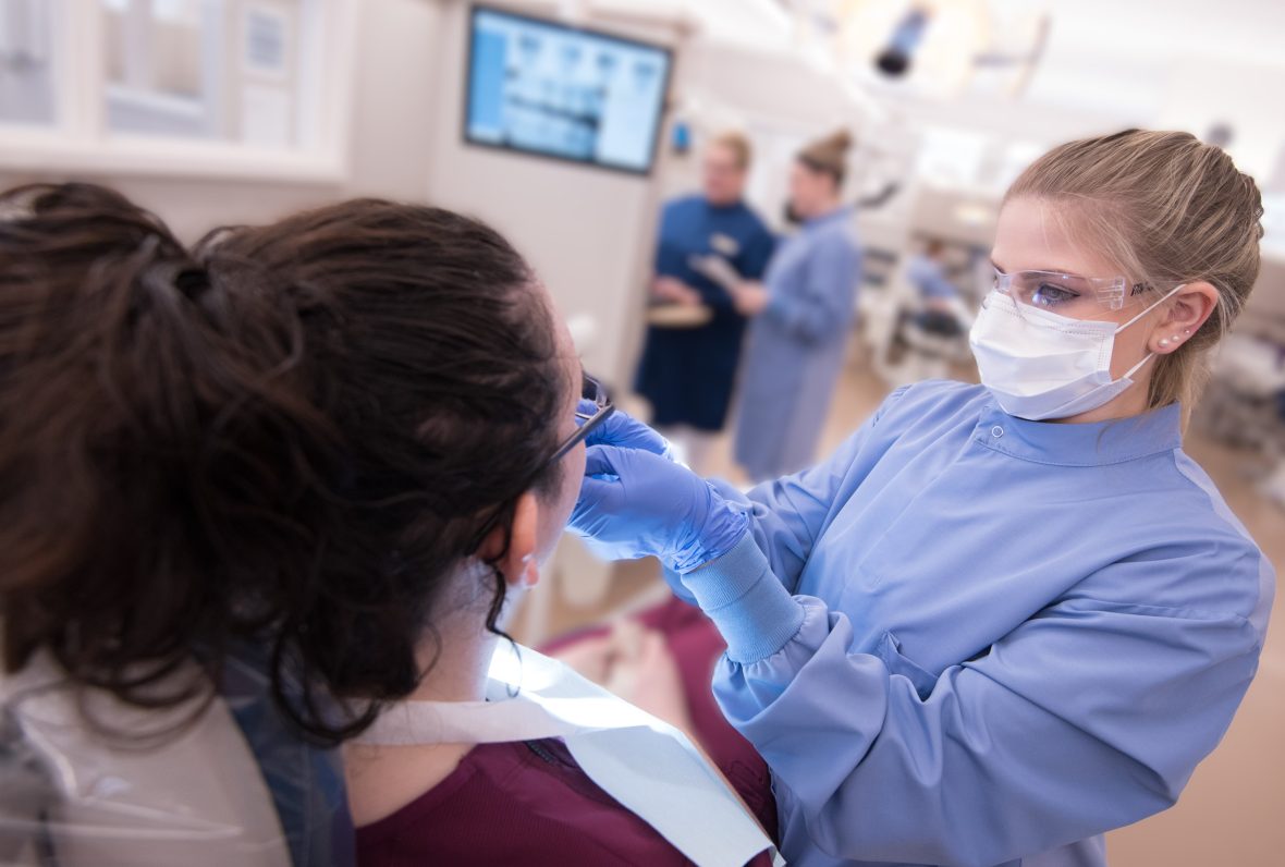 A dental hygiene student wearing protective eyewear, mask, and gloves provides care to a patient in a modern clinic setting, with instructors and equipment visible in the background.