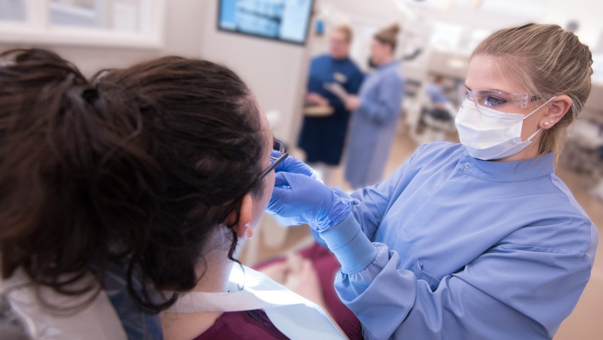 A dental hygiene student wearing protective eyewear, mask, and gloves provides care to a patient in a modern clinic setting, with instructors and equipment visible in the background.