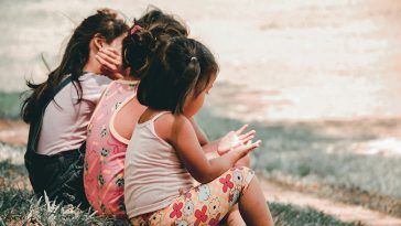 Three young children sit together on the grass outdoors, their backs turned to the camera, sharing a quiet moment.