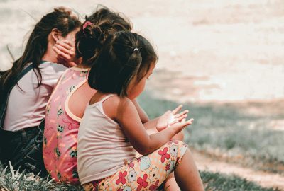 Three young children sit together on the grass outdoors, their backs turned to the camera, sharing a quiet moment.