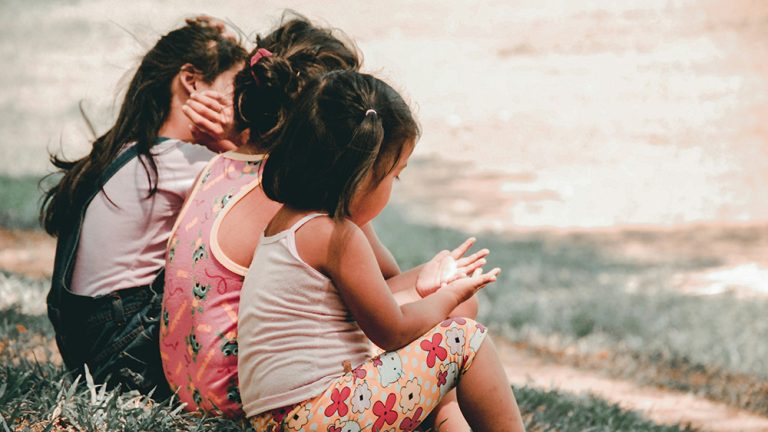 Three young children sit together on the grass outdoors, their backs turned to the camera, sharing a quiet moment.