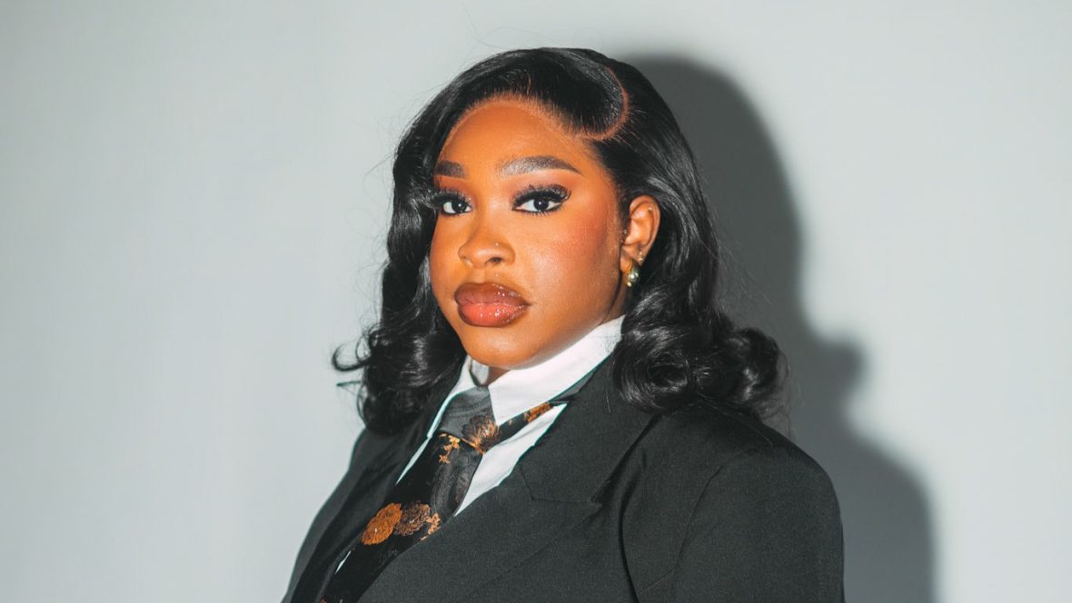 Portrait of Jemima George wearing a black suit and patterned tie, looking confidently toward the camera.