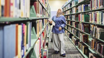 A University of Maine at Augusta student walks through library stacks holding books while browsing titles in a campus library.