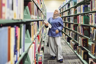 A University of Maine at Augusta student walks through library stacks holding books while browsing titles in a campus library.