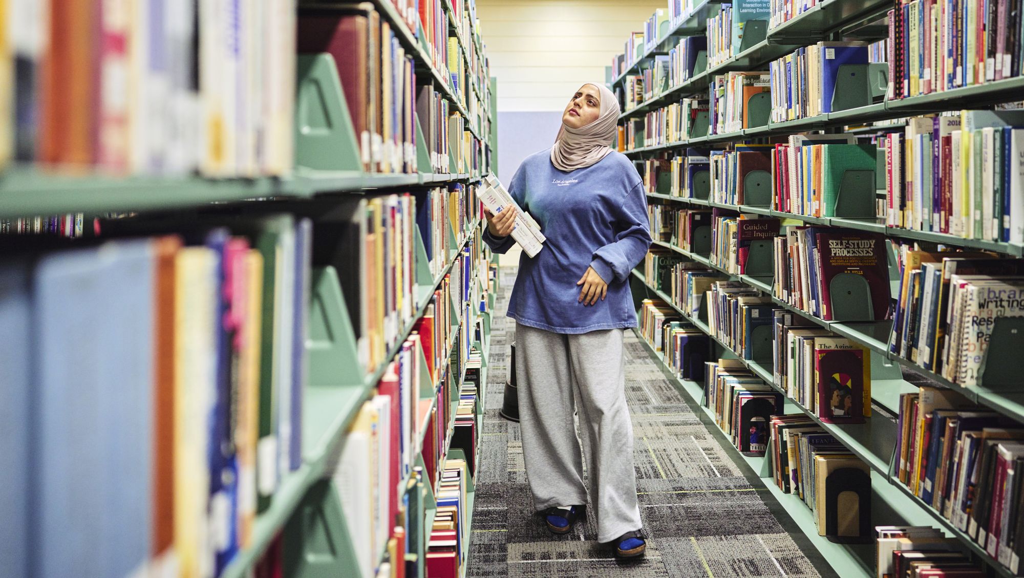 A University of Maine at Augusta student walks through library stacks holding books while browsing titles in a campus library.