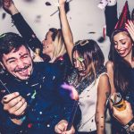 A group of young adults celebrating at a lively party, smiling and laughing as confetti falls around them, some holding party poppers and wearing festive accessories.