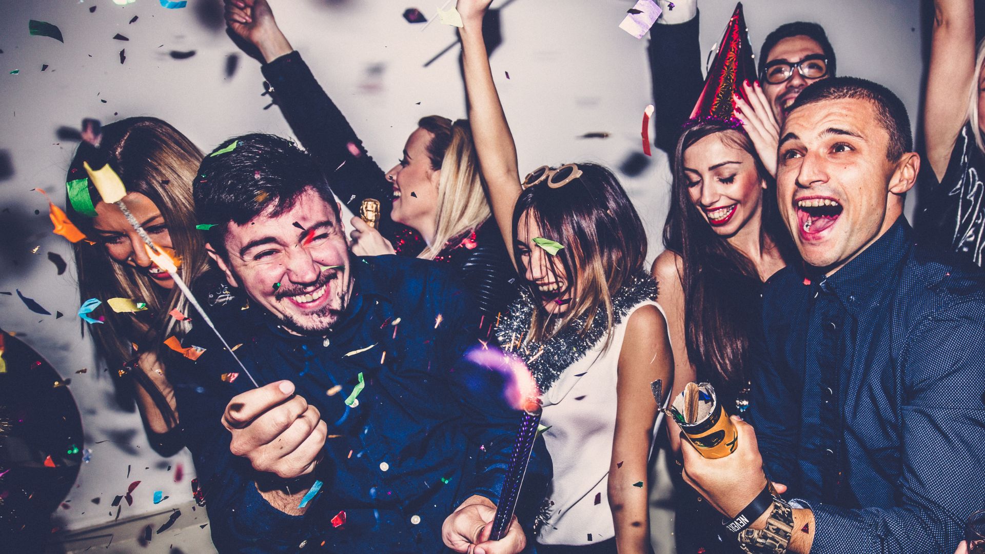 A group of young adults celebrating at a lively party, smiling and laughing as confetti falls around them, some holding party poppers and wearing festive accessories.