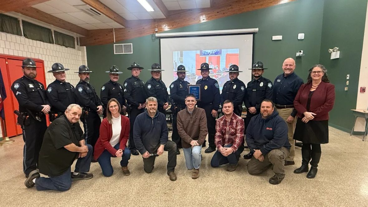 Ten uniformed members of the Winthrop Police Department stand in the back row and 
six members of the Winthrop Town Council kneel in the front row. Chief (ret.) Noel March, and UMA President Jenifer Cushman stand to the right. Two officers in the middle of the back row hold a plaque.
