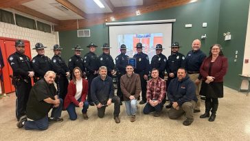 Ten uniformed members of the Winthrop Police Department stand in the back row and six members of the Winthrop Town Council kneel in the front row. Chief (ret.) Noel March, and UMA President Jenifer Cushman stand to the right. Two officers in the middle of the back row hold a plaque.