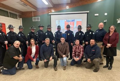 Ten uniformed members of the Winthrop Police Department stand in the back row and six members of the Winthrop Town Council kneel in the front row. Chief (ret.) Noel March, and UMA President Jenifer Cushman stand to the right. Two officers in the middle of the back row hold a plaque.