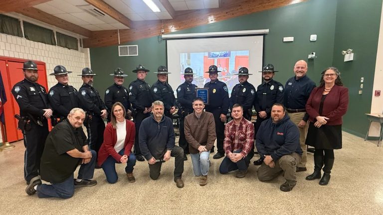 Ten uniformed members of the Winthrop Police Department stand in the back row and six members of the Winthrop Town Council kneel in the front row. Chief (ret.) Noel March, and UMA President Jenifer Cushman stand to the right. Two officers in the middle of the back row hold a plaque.