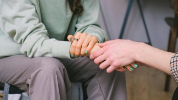 Close-up of seated person between shoulders and knees. Another person holds one of their hands.