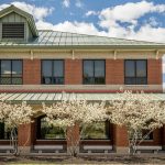 Exterior of a two-story red brick building with a green metal roof, framed by small flowering trees in bloom under a blue sky with scattered clouds.