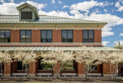 Exterior of a two-story red brick building with a green metal roof, framed by small flowering trees in bloom under a blue sky with scattered clouds.