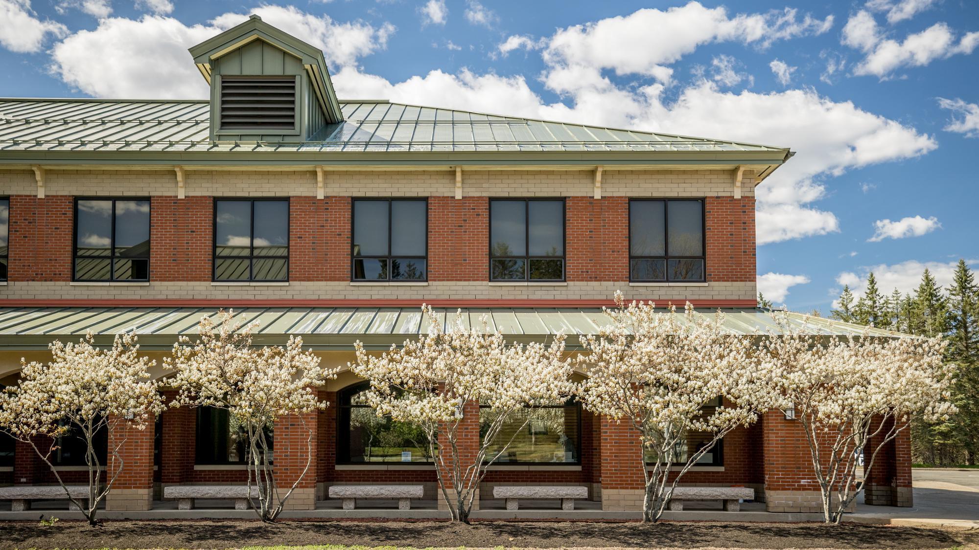 Exterior of a two-story red brick building with a green metal roof, framed by small flowering trees in bloom under a blue sky with scattered clouds.