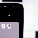 A smartphone screen shows a folder labeled “AI” containing the Gemini and ChatGPT app icons, with a blurred computer screen in the background.