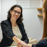 A woman wearing glasses and a blazer smiles and shakes hands with another person across a desk in a small office, suggesting a professional meeting or interview.