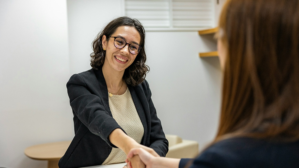 A woman wearing glasses and a blazer smiles and shakes hands with another person across a desk in a small office, suggesting a professional meeting or interview.