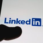 The LinkedIn logo appears on a smartphone screen, with a finger partially visible in the foreground, suggesting someone using the professional networking app.