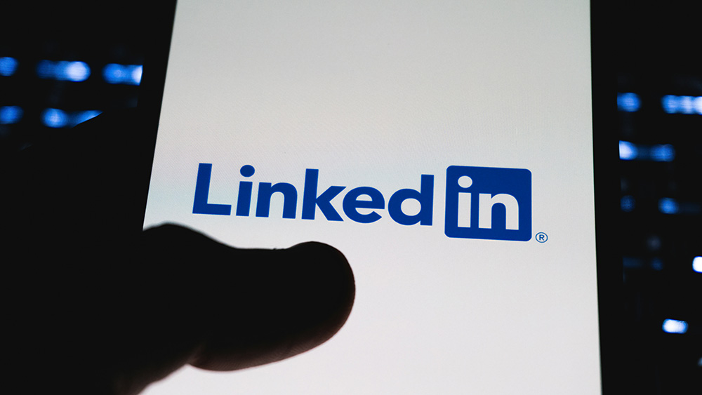 The LinkedIn logo appears on a smartphone screen, with a finger partially visible in the foreground, suggesting someone using the professional networking app.