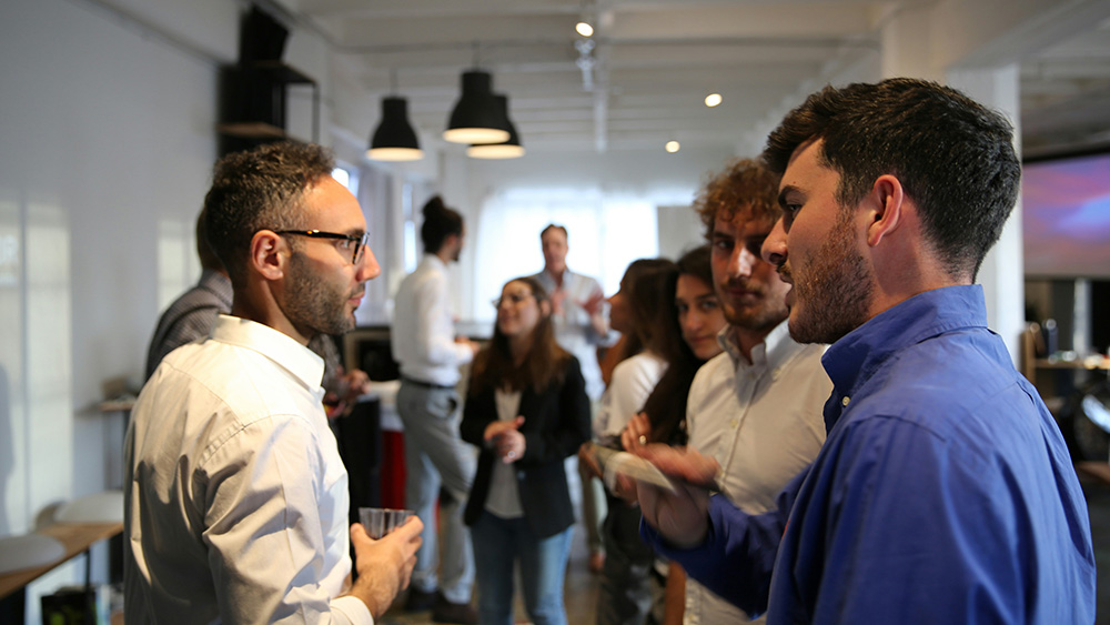 A group of people stand and talk in a bright, modern office space, with several individuals engaged in small conversations, suggesting a professional networking event or informal meeting.