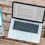 A laptop displaying a document titled “Executive Summary” sits on a wooden table alongside a notebook with a pen, a glass of water, a smartphone, and a small potted plant.
