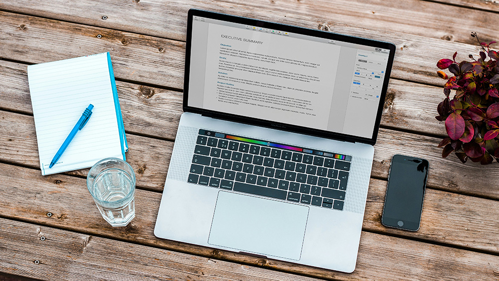A laptop displaying a document titled “Executive Summary” sits on a wooden table alongside a notebook with a pen, a glass of water, a smartphone, and a small potted plant.