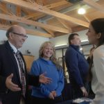 Three adults smile and talk with a young woman at a resource fair table inside a large indoor space. The group appears engaged in conversation, with informational materials visible on the table and other attendees in the background.
