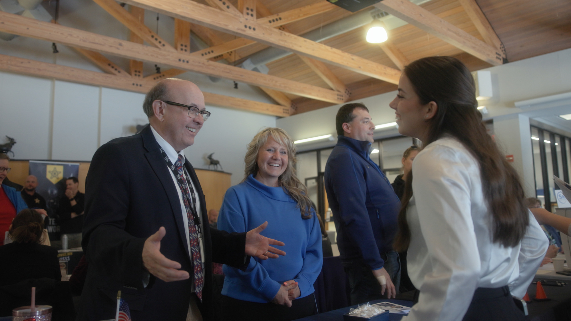 Three adults smile and talk with a young woman at a resource fair table inside a large indoor space. The group appears engaged in conversation, with informational materials visible on the table and other attendees in the background.